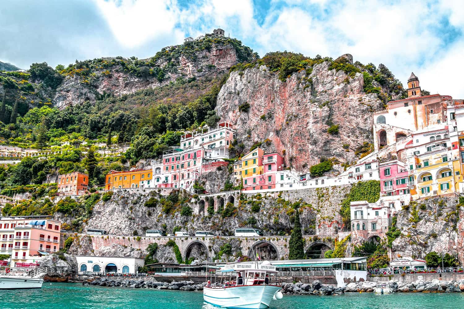 Colorful buildings and lush greenery on a cliffside overlooking the sea in a coastal town, with a boat in the foreground.
