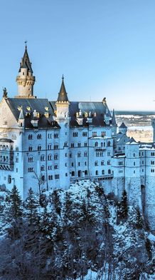 Snow-covered castle on a hill, surrounded by evergreen trees, under a clear blue sky and overlooking a vast landscape.