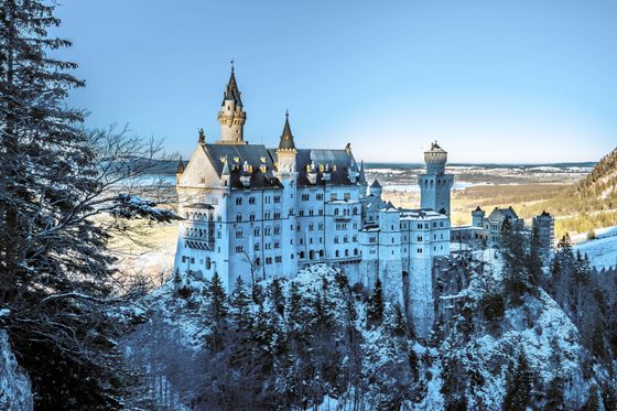 Snow-covered castle on a hill, surrounded by evergreen trees, under a clear blue sky and overlooking a vast landscape.