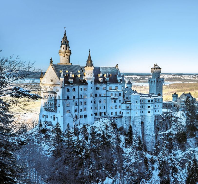 Snow-covered castle on a hill, surrounded by evergreen trees, under a clear blue sky and overlooking a vast landscape.