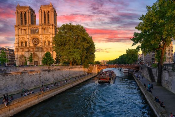 Notre-Dame Cathedral by the Seine River at sunset, with a boat full of people and a colorful sky.