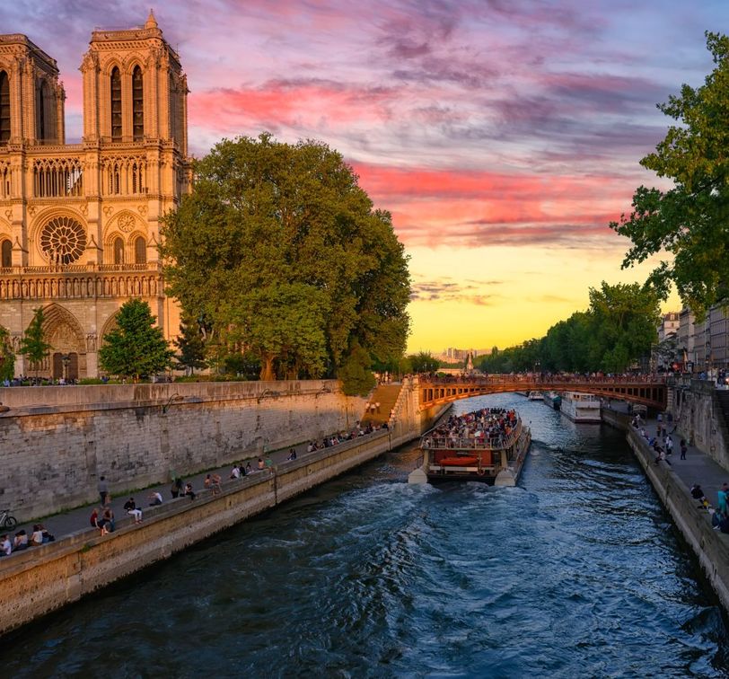 Notre-Dame Cathedral by the Seine River at sunset, with a boat full of people and a colorful sky.