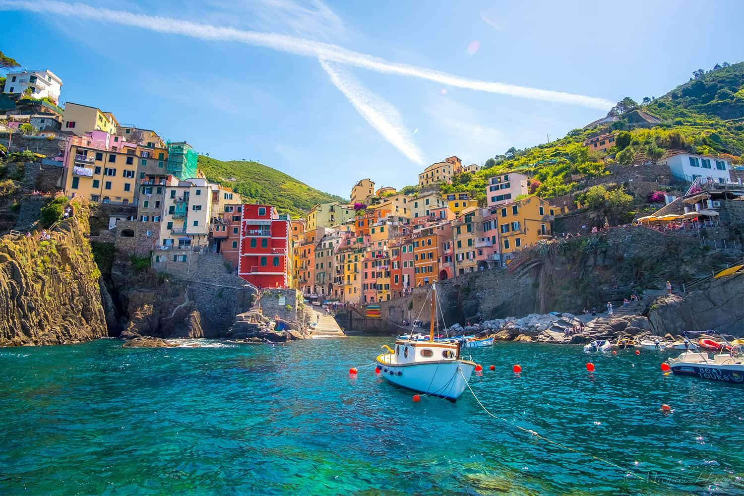 Colorful cliffside buildings overlook a small fishing boat on clear, turquoise waters in the picturesque coastal village under a blue sky.