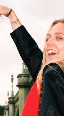 Smiling woman playfully points at Big Ben in the background on a cloudy day, with part of a bridge visible beside her.
