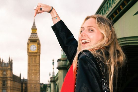 Smiling woman playfully points at Big Ben in the background on a cloudy day, with part of a bridge visible beside her.