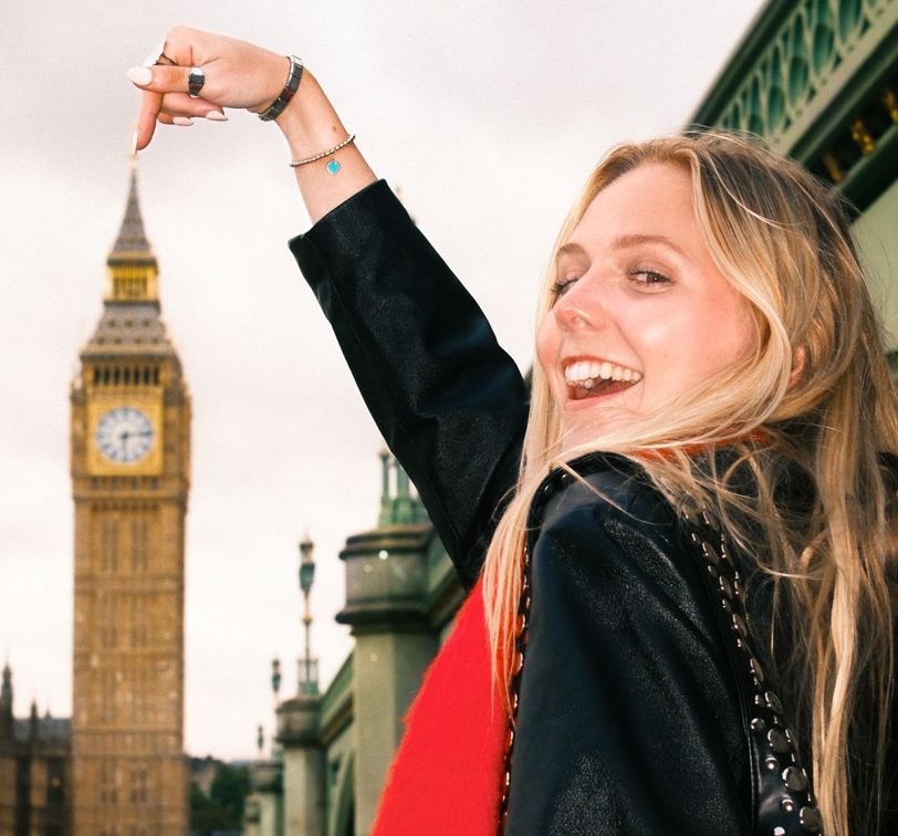 Smiling woman playfully points at Big Ben in the background on a cloudy day, with part of a bridge visible beside her.