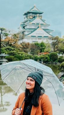 A woman holds a transparent umbrella in a garden, smiling with a Japanese castle in the background.