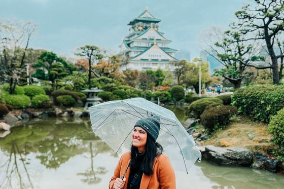 A woman holds a transparent umbrella in a garden, smiling with a Japanese castle in the background.