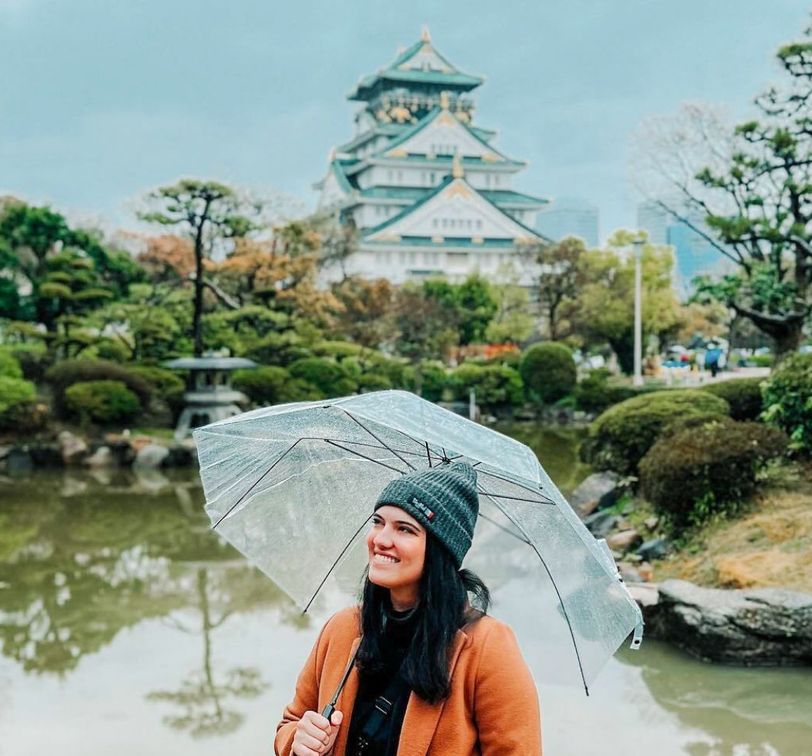 A woman holds a transparent umbrella in a garden, smiling with a Japanese castle in the background.