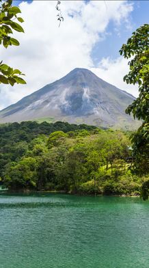 A lush green landscape with a volcano towering in the background, framed by trees, overlooking a serene teal lake under a partly cloudy sky.
