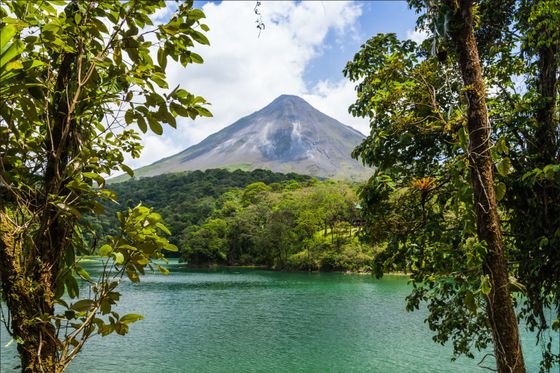 A lush green landscape with a volcano towering in the background, framed by trees, overlooking a serene teal lake under a partly cloudy sky.