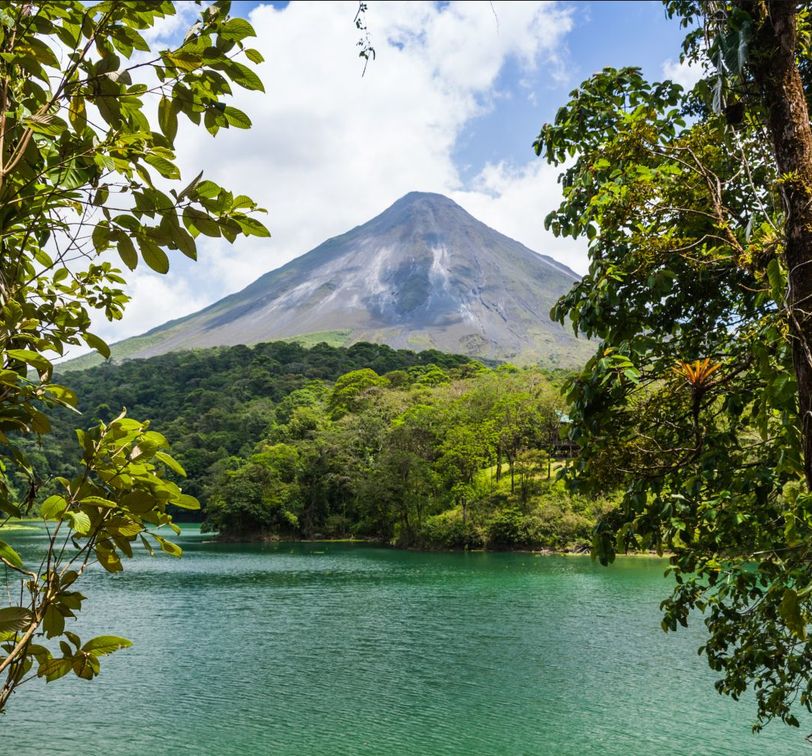 A lush green landscape with a volcano towering in the background, framed by trees, overlooking a serene teal lake under a partly cloudy sky.