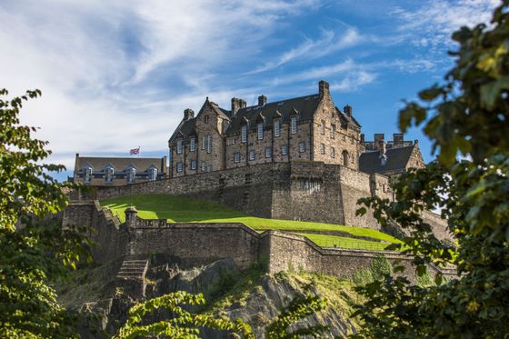 Historic stone castle atop a grassy hill, surrounded by lush greenery and a clear blue sky. A flag flutters above the building.