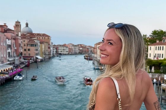 Woman with long hair smiling by a canal filled with boats in Venice, Italy, with historic buildings and a dome in the background at sunset.