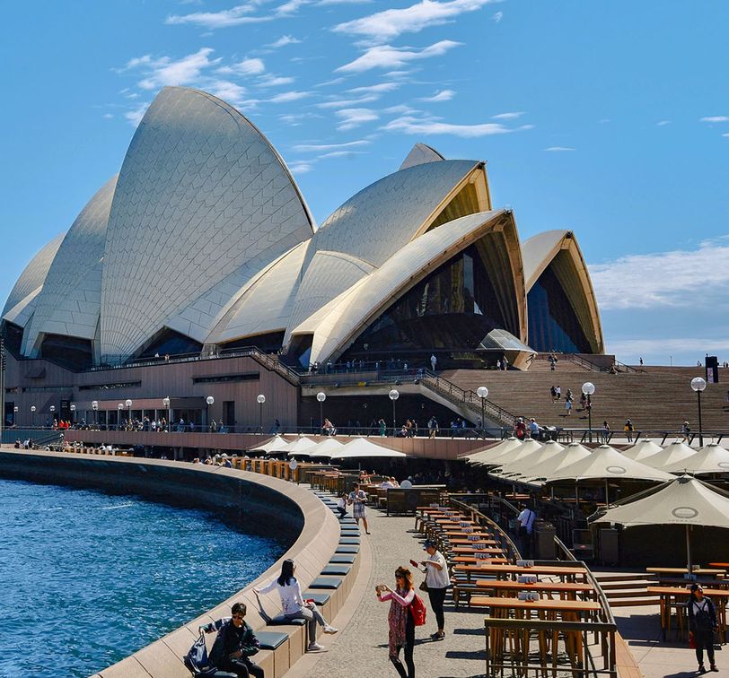 Sydney Opera House with its iconic sails, waterfront promenade, outdoor seating, and people enjoying a sunny day.