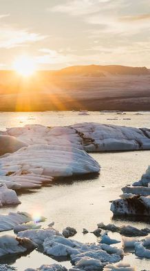 Sunrise over an icy lagoon with large floating icebergs and a distant glacier, under a partly cloudy sky.