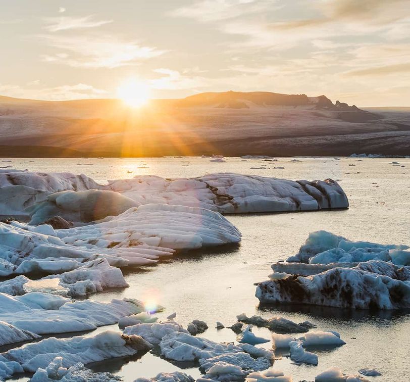 Sunrise over an icy lagoon with large floating icebergs and a distant glacier, under a partly cloudy sky.