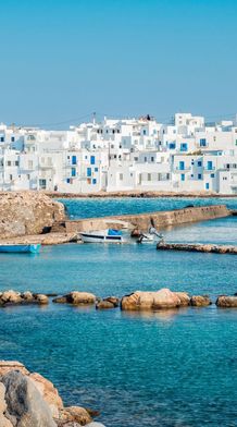 A Greek island harbor with white buildings, blue shutters, and domed churches. Boats are docked in clear turquoise waters with rocky breakwaters.
