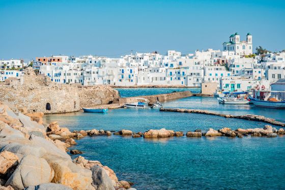 A Greek island harbor with white buildings, blue shutters, and domed churches. Boats are docked in clear turquoise waters with rocky breakwaters.