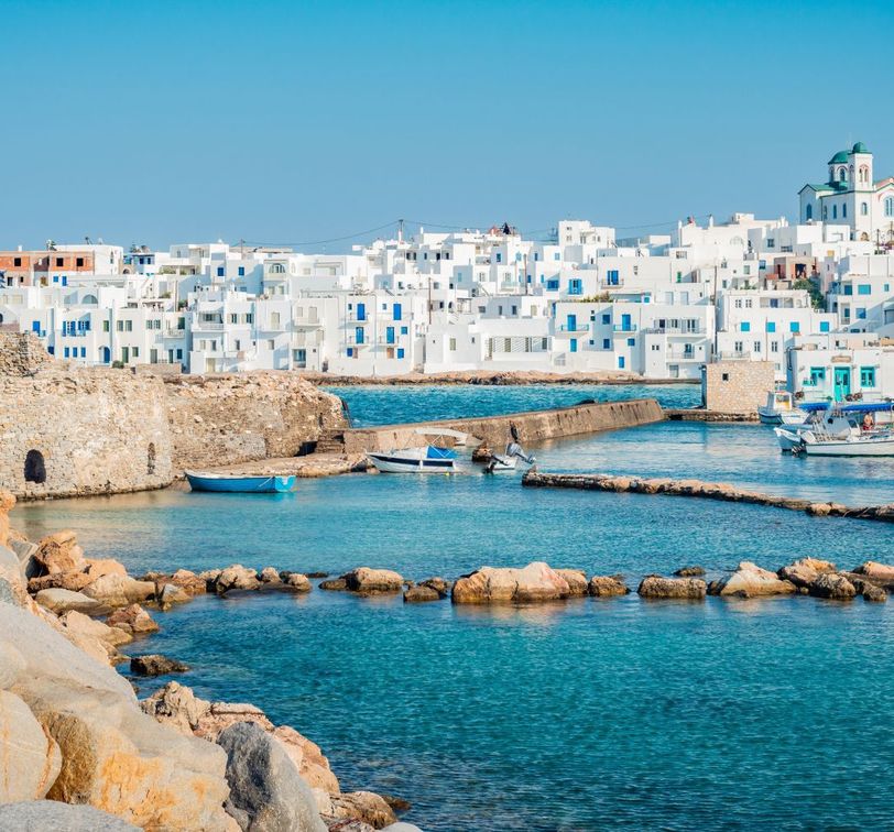 A Greek island harbor with white buildings, blue shutters, and domed churches. Boats are docked in clear turquoise waters with rocky breakwaters.