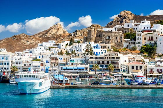 A picturesque Greek seaside town with white buildings, a mountain backdrop, a harbor with boats, and clear blue water under a sunny sky.