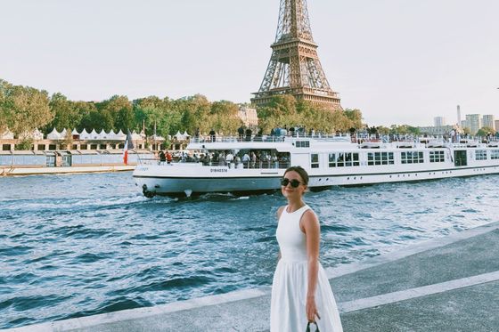 A woman in a white dress stands by the Seine River with a boat and the Eiffel Tower in the background on a sunny day.