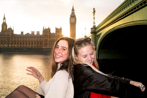Two smiling women pose back-to-back on a bridge with Big Ben and the Houses of Parliament in the background at sunset.