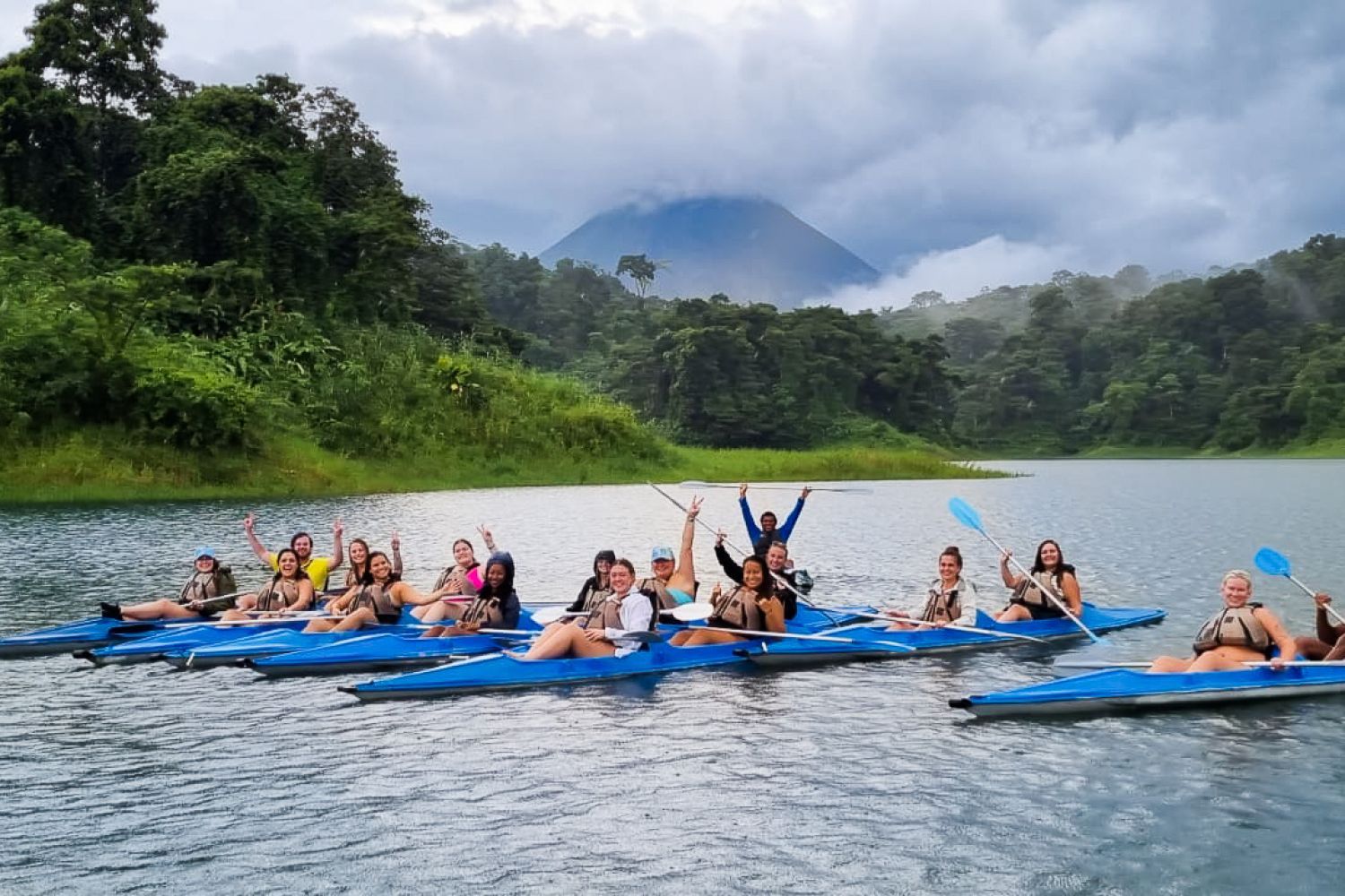 Group of people kayaking on a lake surrounded by lush greenery and mountains under a cloudy sky.