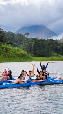 Group of people kayaking on a lake surrounded by lush greenery and mountains under a cloudy sky.