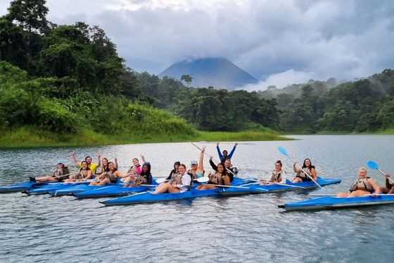 Group of people kayaking on a lake surrounded by lush greenery and mountains under a cloudy sky.