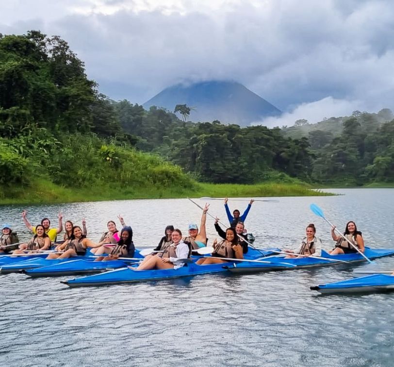 Group of people kayaking on a lake surrounded by lush greenery and mountains under a cloudy sky.