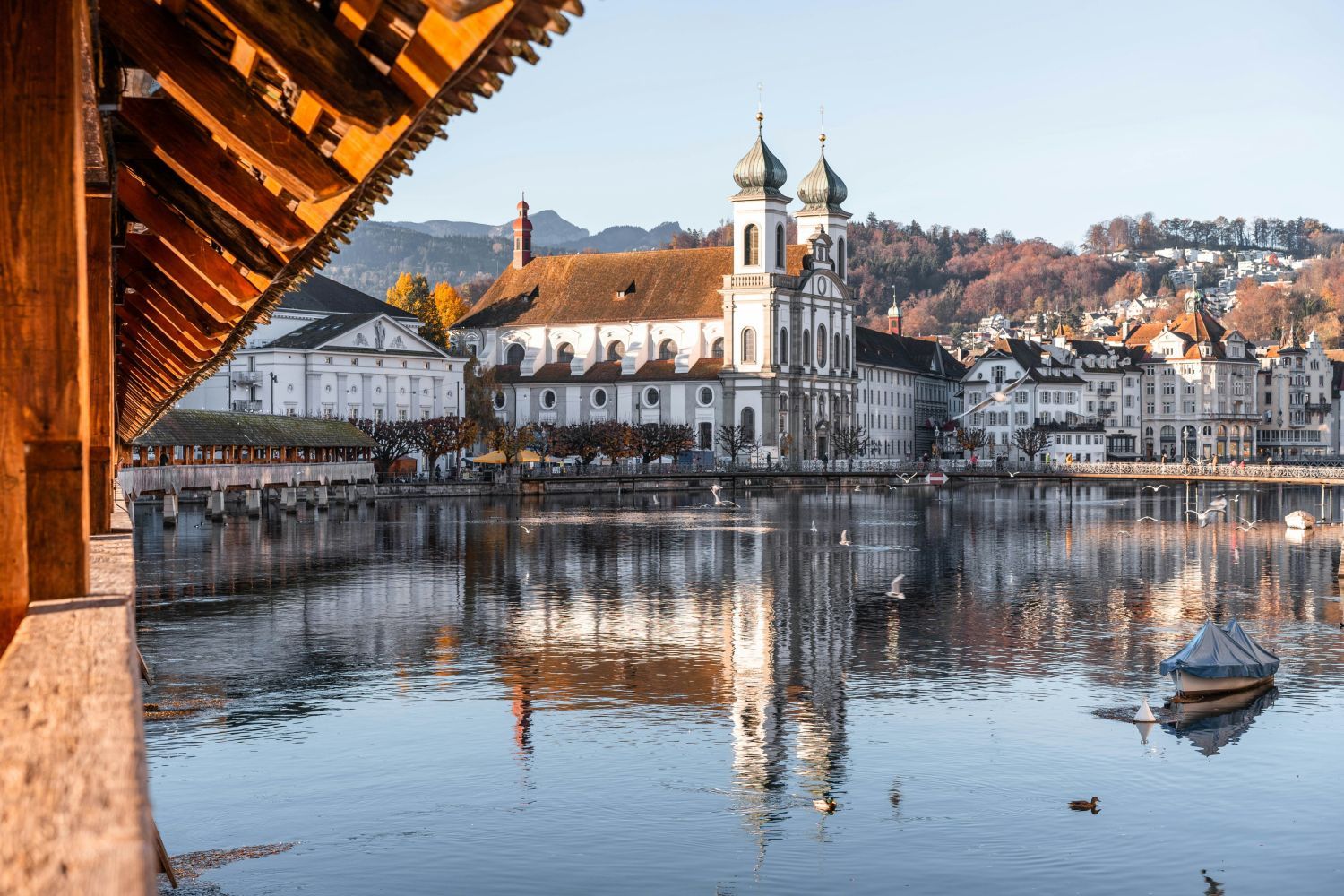 Historic wooden bridge over a calm river, with a white church and Alps in the background, under a clear blue sky.