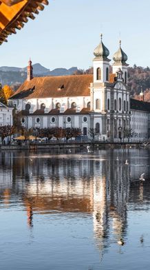 Historic wooden bridge over a calm river, with a white church and Alps in the background, under a clear blue sky.