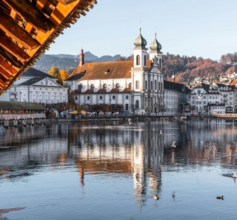 Historic wooden bridge over a calm river, with a white church and Alps in the background, under a clear blue sky.