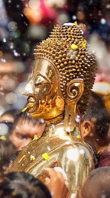 Golden Buddha statue being carried through a crowd during a water festival, with water splashes and petals in the air.