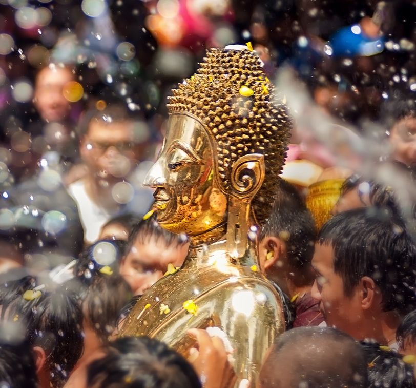 Golden Buddha statue being carried through a crowd during a water festival, with water splashes and petals in the air.