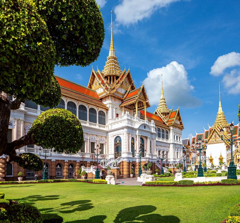 A grand palace with ornate, golden spires and white facades, surrounded by lush, manicured gardens under a bright blue sky with fluffy clouds.