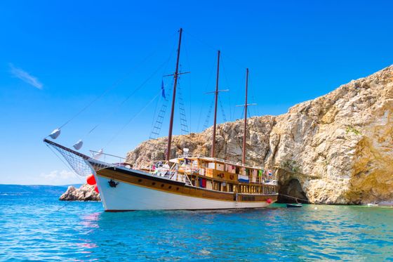 A large wooden sailboat anchored near a rocky cliff in clear blue water with a bright, sunny sky.