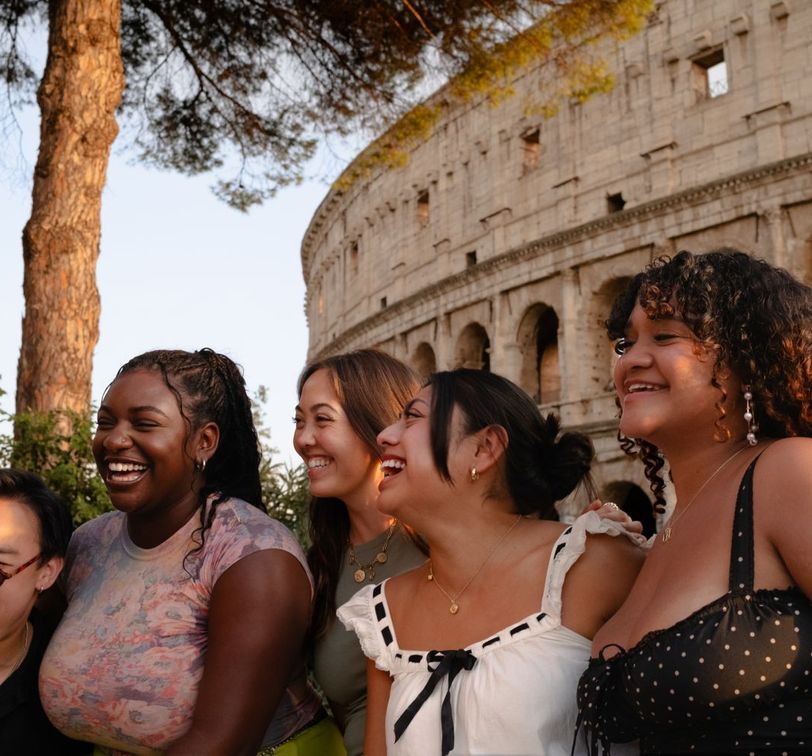Five people smiling near the Colosseum at sunset, with trees and the ancient structure in the background.
