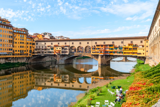 Ponte Vecchio bridge over the Arno River in Florence, Italy, with colorful buildings, clear blue sky, and people relaxing on the grassy riverbank.