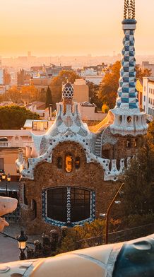 Sunset view of Park Güell in Barcelona, featuring colorful mosaic architecture, organic shapes, and a panoramic cityscape in the background.