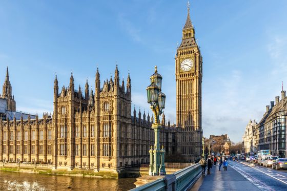 The Elizabeth Tower and the Palace of Westminster in London with people walking on Westminster Bridge under a clear blue sky.