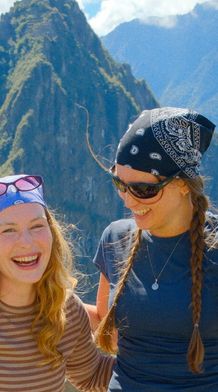 Two smiling women wearing bandanas and sunglasses stand in front of Machu Picchu, with mountains in the background on a sunny day.