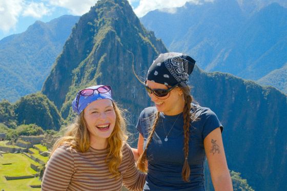 Two smiling women wearing bandanas and sunglasses stand in front of Machu Picchu, with mountains in the background on a sunny day.