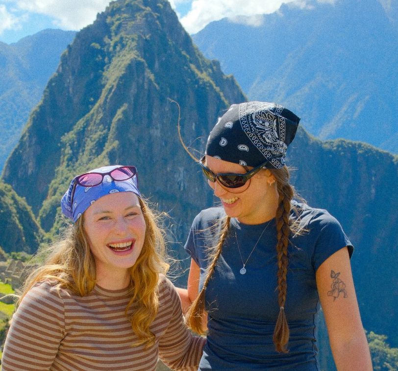 Two smiling women wearing bandanas and sunglasses stand in front of Machu Picchu, with mountains in the background on a sunny day.