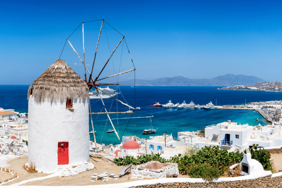 A white traditional windmill with a thatched roof overlooks a vibrant blue sea, boats, and white buildings on a sunny day in Mykonos, Greece.