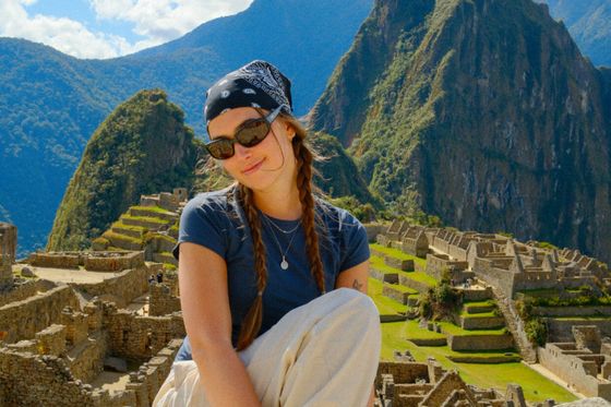 A woman with sunglasses and a bandana sits in front of the ancient ruins of Machu Picchu, surrounded by green mountains under a blue sky.