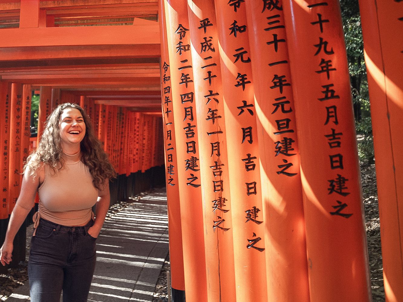 A woman smiles while walking through a tunnel of orange torii gates with Japanese inscriptions in bright sunlight.