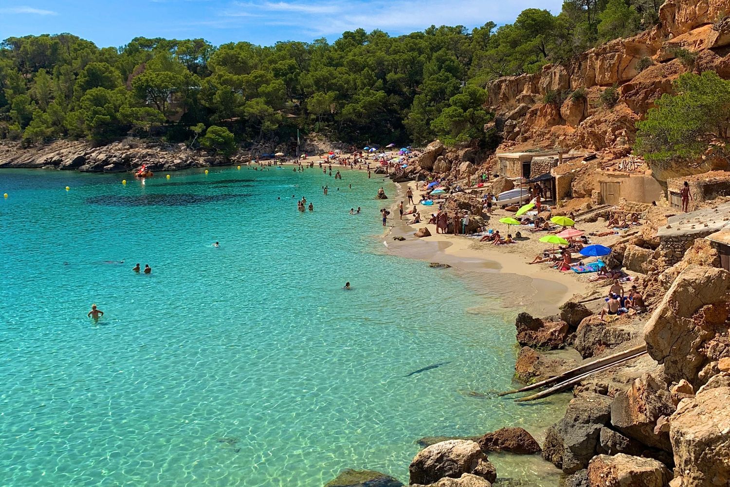 A rocky beach with clear turquoise water, people swimming and sunbathing on sandy shores, surrounded by green trees and cliffs.