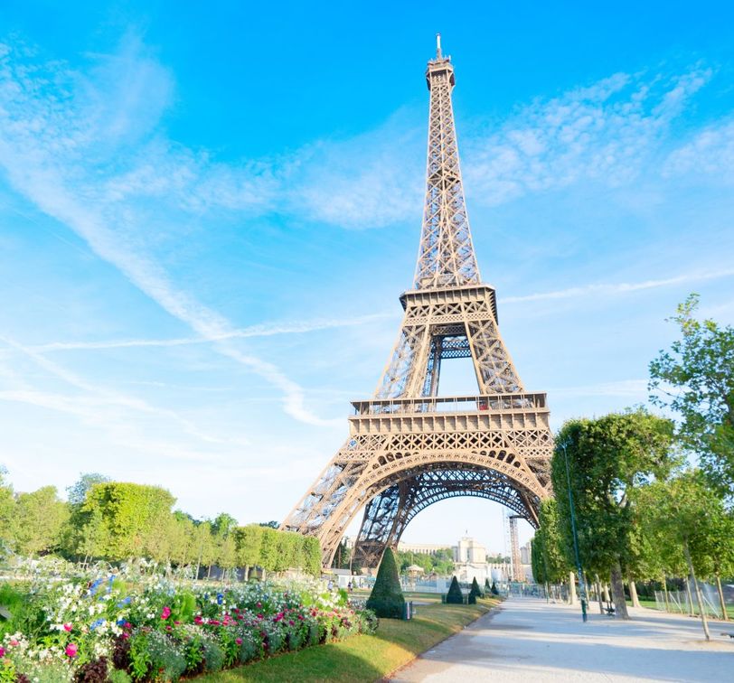 The Eiffel Tower surrounded by green trees and colorful flowers under a clear blue sky.